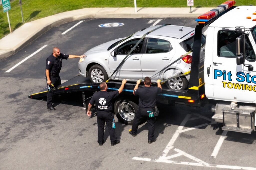 Team of tow truck operators securing a silver vehicle onto a flatbed for impound towing in a parking lot.
